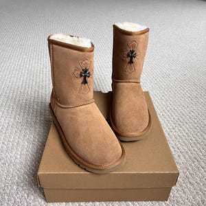 May include: A pair of tan suede boots with a cross design. The boots have a short shaft, a round toe, and a brown sole. The cross design is black and gold. The boots are sitting on a brown cardboard box.
