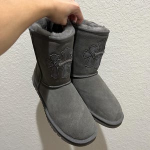 May include: A pair of gray suede boots with a cross design on the front. The boots have a soft, furry lining and a rubber sole. The boots are held up by a hand.
