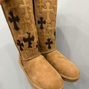 May include: A pair of tan suede mid-calf boots featuring a thick, light-colored interior lining. The exterior of the boots is decorated with numerous stitched cross-shaped patches in varying shades of tan, beige, and dark brown leather.