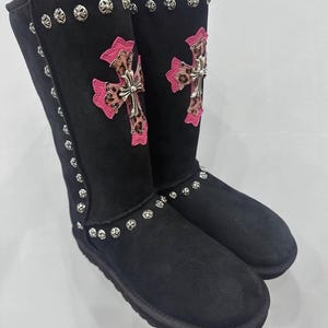 May include: A pair of black mid-calf boots featuring silver-toned circular metal studs along the seams. Each boot displays a prominent cross emblem with a pink border and a leopard print center, accented by a metallic silver cross overlay.