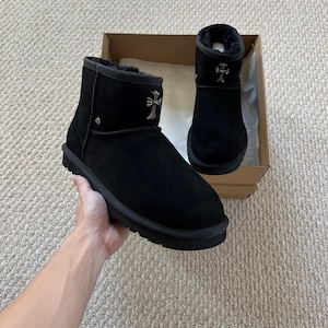 May include: Black suede ankle boots with a silver cross emblem on the front. The boots have a thick, black sole and a soft, fur-like lining. The boots are displayed in a cardboard box.