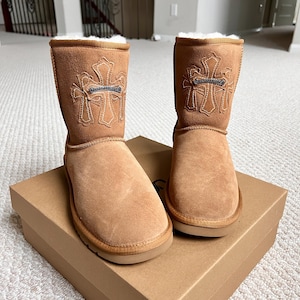 May include: A pair of tan suede boots with a cross design on the side. The boots have a soft, fluffy lining and are sitting on a cardboard box. The boots are a neutral color and appear to be new.