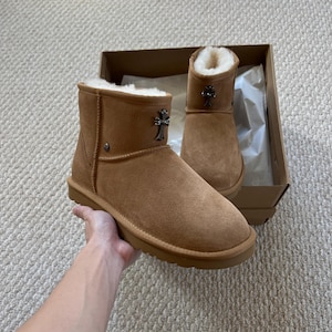May include: A pair of brown suede ankle boots with a cross emblem. The boots have a soft, furry interior and a rubber sole. One boot is held in a hand, and the other is in a cardboard box.
