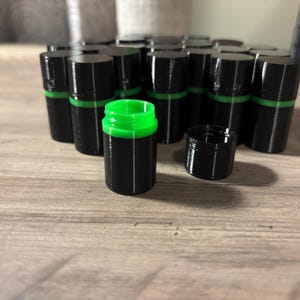 May include: A collection of small, cylindrical black containers with bright green inner rims and matching black screw-on lids. One container is shown open in the foreground, revealing the vibrant green interior, while the others remain closed.