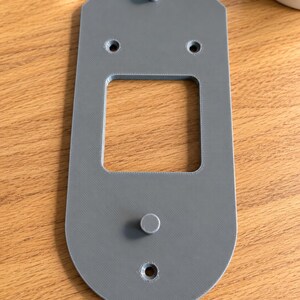 May include: A gray, elongated, flat mounting plate with rounded ends, featuring a central square cutout, two small cylindrical pegs, and three countersunk screw holes arranged vertically along the center line of the object.