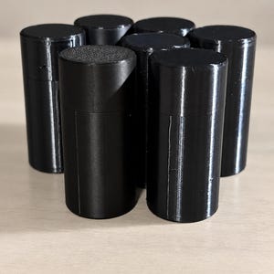 May include: A collection of seven black cylindrical containers arranged in a cluster. Each container features a smooth, vertical body with a matching flat circular lid on top, creating a uniform set of small, upright storage vessels.