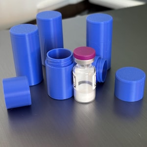 May include: A clear glass vial with a pink cap and white powder inside, surrounded by several blue cylindrical containers with lids. The containers are arranged on a gray surface.