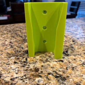 May include: A bright neon green 3D-printed object with three circular holes. The object has a rectangular shape with two supporting legs at the bottom. The item is sitting on a speckled countertop.