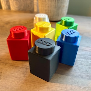 May include: A collection of six plastic containers shaped like large building blocks in red, yellow, white, green, blue, and black. Each block features a circular lid with the embossed LEGO logo on top, creating a stackable storage set.