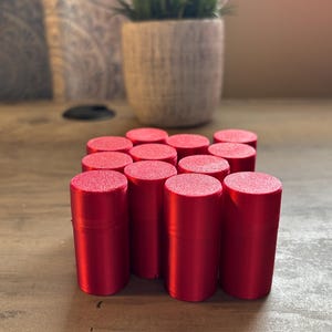 May include: A collection of twelve cylindrical red containers arranged in a grid. Each container features a smooth, solid red body with a textured, circular lid on top. The items are uniform in size and shape, standing upright in a cluster.
