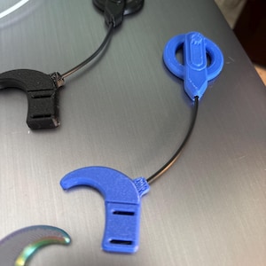May include: Several small, 3D-printed devices in black and blue are displayed. Each device has a curved, hook-like shape connected to a circular or oval component by a thin, black wire. The blue devices have a lighter, brighter appearance.
