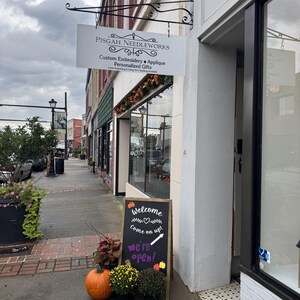Puede incluir: Fachada de una tienda con un letrero que dice "PISGAH NEEDLEWORKS". Un letrero de pizarra dice "&iexcl;Bienvenido, entra! &iexcl;Estamos abiertos!" con una calabaza y crisantemos en macetas.