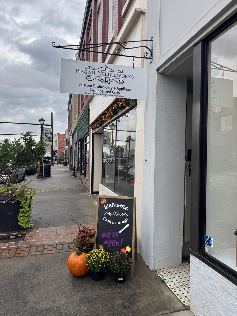 Puede incluir: Fachada de una tienda con un letrero que dice "PISGAH NEEDLEWORKS". Un letrero de pizarra dice "&iexcl;Bienvenido, entra! &iexcl;Estamos abiertos!" con una calabaza y crisantemos en macetas.