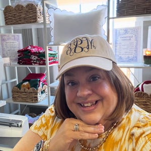 Puede incluir: Gorra de b&eacute;isbol beige con bordado de monograma azul marino. La gorra la lleva una persona. Estantes en el fondo exhiben almohadas, cestas y otros art&iacute;culos.
