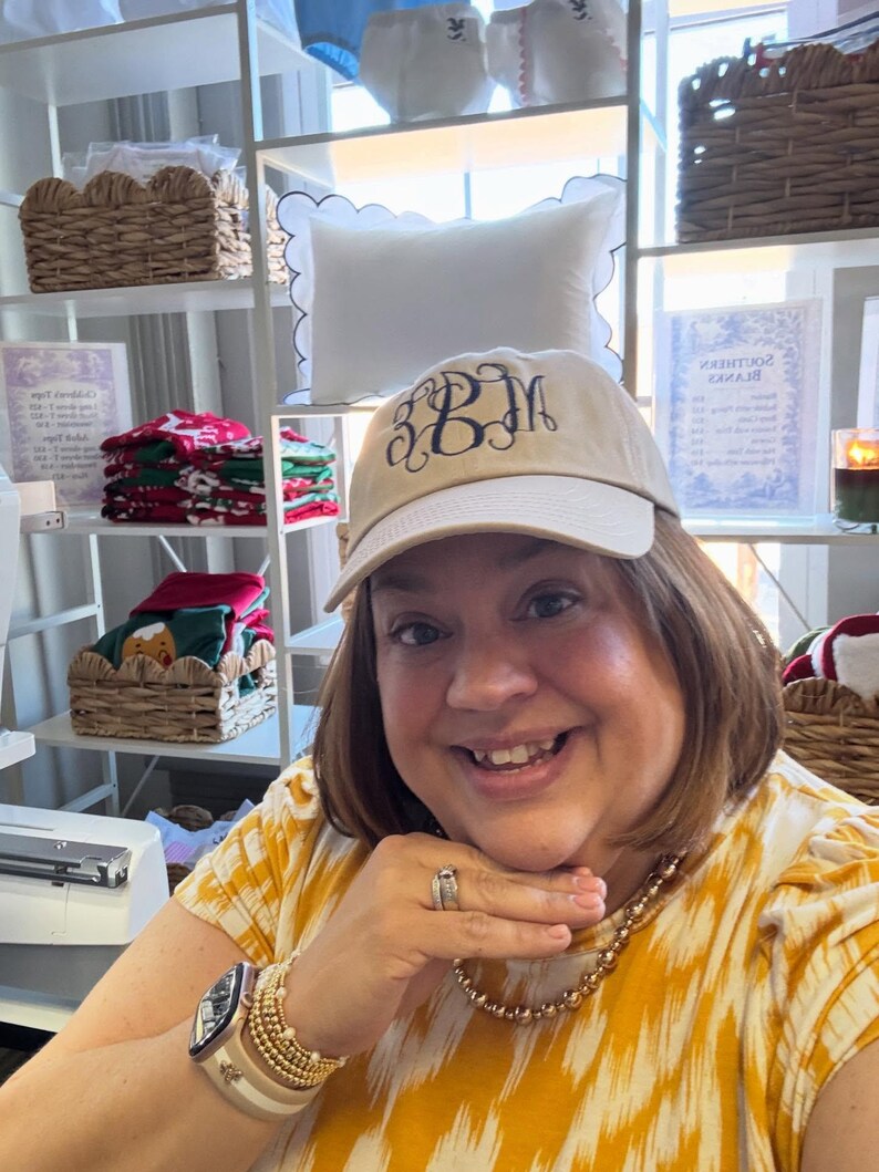 Puede incluir: Gorra de b&eacute;isbol beige con bordado de monograma azul marino. La gorra la lleva una persona. Estantes en el fondo exhiben almohadas, cestas y otros art&iacute;culos.