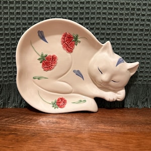 May include: A cream-colored ceramic trinket dish shaped like a sleeping cat. The dish features a glossy finish with a subtle crackle texture, decorated with three red carnation flowers, green stems, and small blue petal accents.