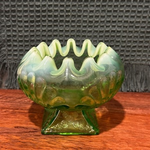May include: A green glass bowl with a ruffled rim and a square base. The bowl has a decorative pattern on the base and a wavy edge. The glass has a slight iridescence, giving it a vintage look.