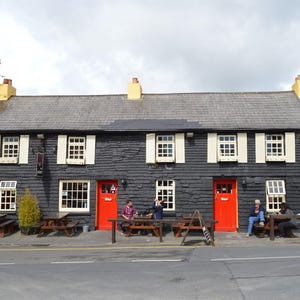 May include: A two-story building with black horizontal siding, a gray slate roof, and two bright red doors labeled Bar. The exterior features white window frames with cream shutters, three yellow chimney stacks, and wooden picnic tables.