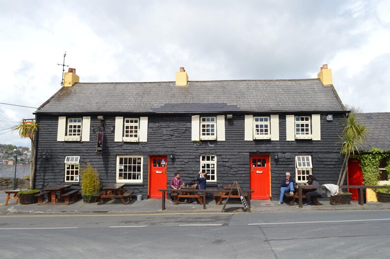 May include: A two-story building with black horizontal siding, a gray slate roof, and two bright red doors labeled Bar. The exterior features white window frames with cream shutters, three yellow chimney stacks, and wooden picnic tables.