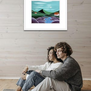 May include: A framed landscape painting featuring a winding blue river flowing through green hills, large purple boulders, and two small white houses with red roofs under a light blue sky with a pale moon and thin white clouds.