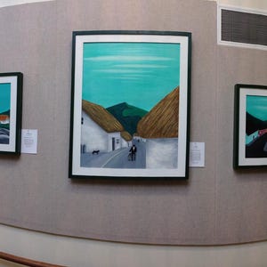May include: A gallery hallway displaying framed landscape paintings. The artworks depict scenes with turquoise skies, green hills, and white buildings. The frames are black, and the paintings are mounted on a beige wall.