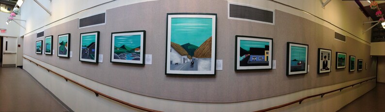 May include: A gallery hallway displaying framed landscape paintings. The artworks depict scenes with turquoise skies, green hills, and white buildings. The frames are black, and the paintings are mounted on a beige wall.