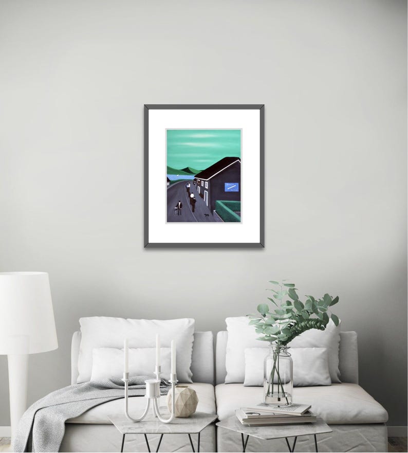 May include: A framed art print featuring a landscape painting of a dark building with a blue sign, a road with figures, and green hills under a light green sky. The artwork is enclosed in a thin black frame with a white mat border.