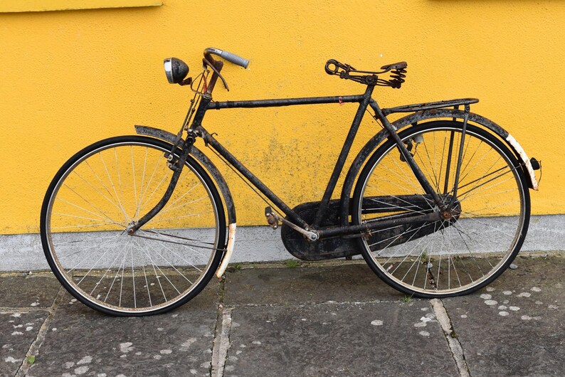 May include: A vintage black bicycle featuring a metal frame, a sprung leather saddle, a chain guard, and a front headlight. The bike includes thin tires, wire-spoked wheels, and white-tipped fenders, showcasing a weathered, aged appearance.