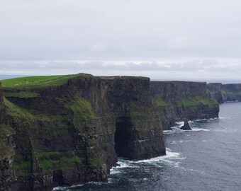 May include: The Cliffs of Moher feature steep, dark brown rock faces topped with vibrant green grass, extending into the deep blue Atlantic Ocean under a cloudy, overcast sky, with small white waves crashing against the base of the cliffs.