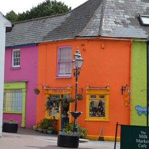 May include: A street scene featuring a row of vibrant buildings painted in bright pink, orange, and teal. A black metal street lamp stands in the center. A sign on the teal building reads The Milk Market, and a green sign reads The Milk Market Cafe.