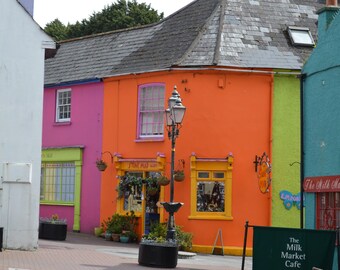 May include: A street scene featuring a row of vibrant buildings painted in bright pink, orange, and teal. A black metal street lamp stands in the center. A sign on the teal building reads The Milk Market, and a green sign reads The Milk Market Cafe.