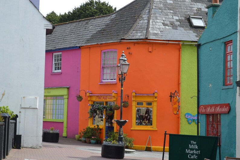 May include: A street scene featuring a row of vibrant buildings painted in bright pink, orange, and teal. A black metal street lamp stands in the center. A sign on the teal building reads The Milk Market, and a green sign reads The Milk Market Cafe.