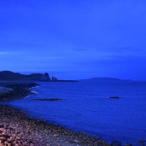 May include: A coastal landscape at twilight featuring a rocky shoreline meeting a deep blue sea under a dark sky. The horizon shows distant hills and a small structure on the headland, all bathed in a monochromatic blue light.