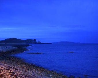 May include: A coastal landscape at twilight featuring a rocky shoreline meeting a deep blue sea under a dark sky. The horizon shows distant hills and a small structure on the headland, all bathed in a monochromatic blue light.