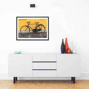 May include: A framed wall art print featuring a black bicycle parked against a vibrant yellow wall. The bicycle has a classic frame, thin wheels, and a rear rack. The print is displayed in a thin black frame, showcasing a high-contrast scene with a bright yellow background and a dark gray pavement surface.