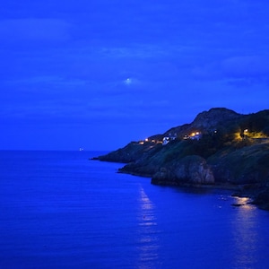 May include: A coastal landscape at twilight featuring a deep blue ocean meeting a rocky shoreline. A line of warm yellow lights from houses and street lamps illuminates the cliffside, while a faint moon is visible in the dark blue sky.