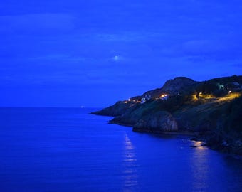 Könnte beinhalten: Eine Küstenlandschaft in der Dämmerung mit einem tiefblauen Ozean und einer felsigen Küste. Eine Reihe warmer gelber Lichter von Häusern und Straßenlaternen beleuchtet den Hang, während ein blasser Mond am dunklen Himmel steht.