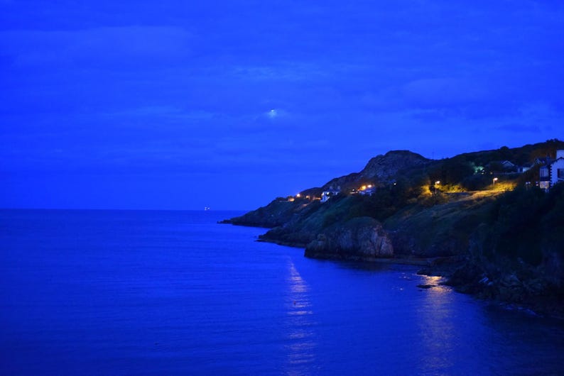 May include: A coastal landscape at twilight featuring a deep blue ocean meeting a rocky shoreline. A line of warm yellow lights from houses and street lamps illuminates the cliffside, while a faint moon is visible in the dark blue sky.