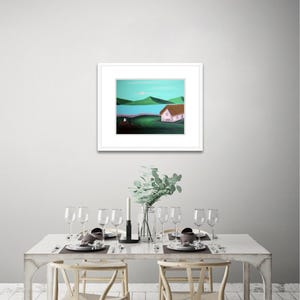May include: A framed landscape painting featuring a white cottage with a brown roof, situated on a dark green field. In the background, a bright blue body of water is bordered by rolling green hills under a light blue sky.