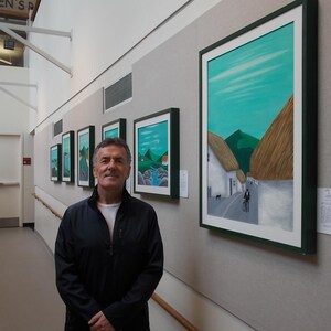 May include: Several framed landscape paintings with green and white mounts are displayed on a grey wall. The artworks show scenes with green hills, blue skies, and white buildings. A man in a black jacket and jeans stands in front of the paintings.