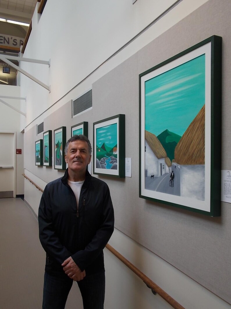 May include: Several framed landscape paintings with green and white mounts are displayed on a grey wall. The artworks show scenes with green hills, blue skies, and white buildings. A man in a black jacket and jeans stands in front of the paintings.