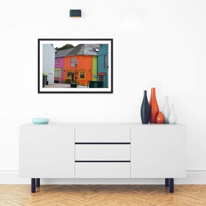 May include: A framed wall art print featuring a colorful street scene with bright pink, orange, and teal buildings. The image shows a storefront with a sign that reads The Milk Market Cafe. The print is displayed above a white sideboard.