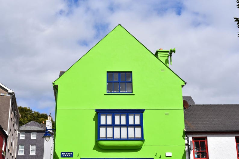 May include: A bright lime green building facade featuring a prominent blue bay window on the lower level and a single rectangular blue window above. A small blue street sign with white text reads Market Lane on the corner of the structure.