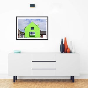 May include: A framed wall art print featuring a bright lime green house with a dark blue window frame and a gabled roof, set against a cloudy blue sky. The artwork is displayed in a black frame hanging above a white sideboard with four vases.