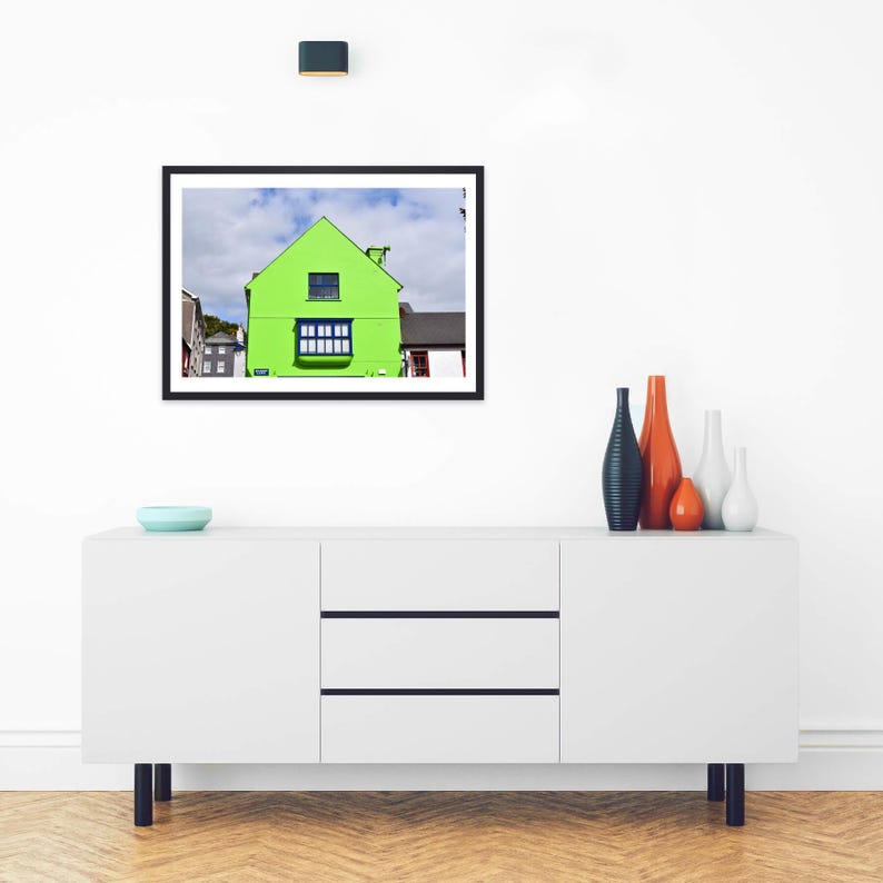 May include: A framed wall art print featuring a bright lime green house with a dark blue window frame and a gabled roof, set against a cloudy blue sky. The artwork is displayed in a black frame hanging above a white sideboard with four vases.