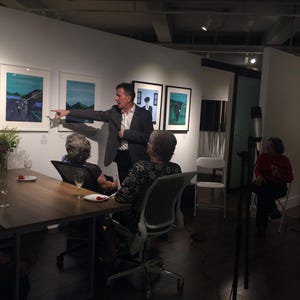 May include: An art gallery event with a speaker gesturing towards framed artwork. Attendees are seated at a table with drinks and flowers. The room is well-lit with spotlights and features modern art.
