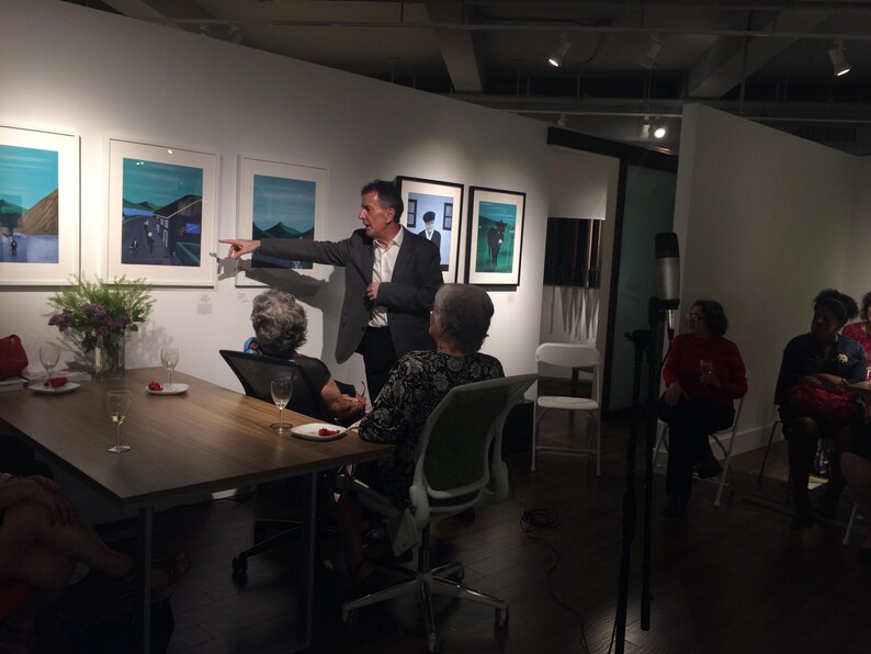May include: An art gallery event with a speaker gesturing towards framed artwork. Attendees are seated at a table with drinks and flowers. The room is well-lit with spotlights and features modern art.