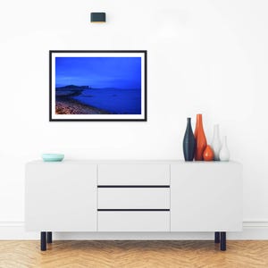 May include: A framed landscape photograph featuring a deep blue twilight seascape with a rocky shoreline and distant hills. The artwork is displayed above a white sideboard cabinet with three drawers and four black legs, accented by four vases.