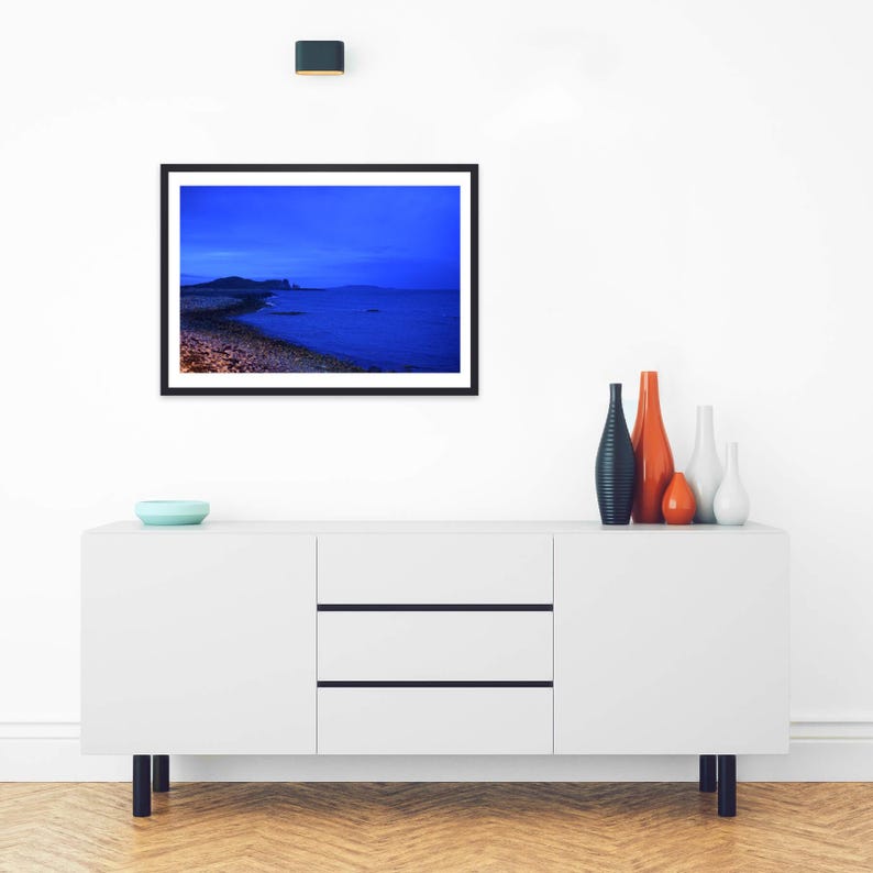 May include: A framed landscape photograph featuring a deep blue twilight seascape with a rocky shoreline and distant hills. The artwork is displayed above a white sideboard cabinet with three drawers and four black legs, accented by four vases.