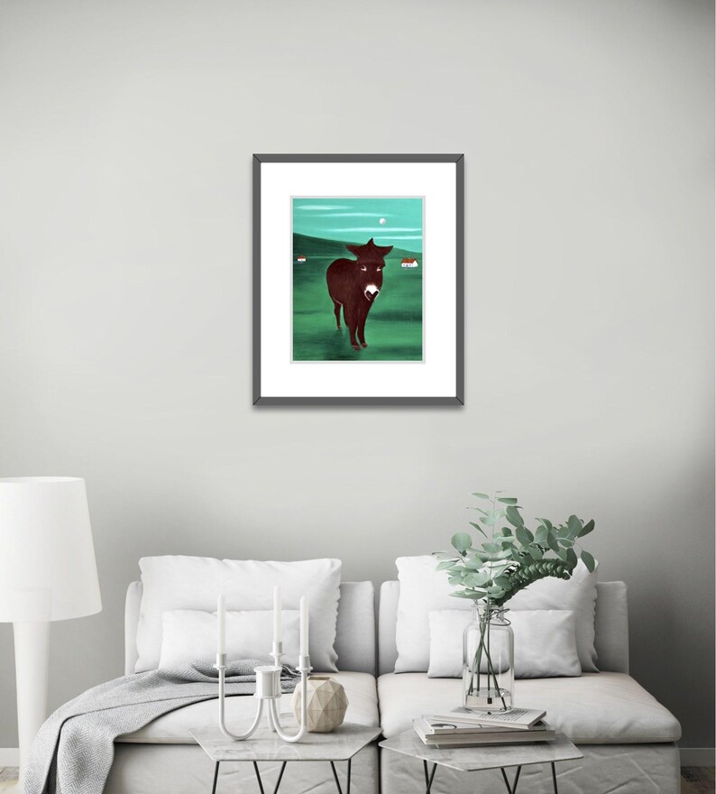 Puede incluir: Una l&aacute;mina de arte enmarcada que muestra un burro de color marr&oacute;n oscuro de pie en un campo verde bajo un cielo verde azulado con una peque&ntilde;a luna blanca y dos edificios blancos distantes. La obra tiene un marco negro fino con paspart&uacute; blanco.